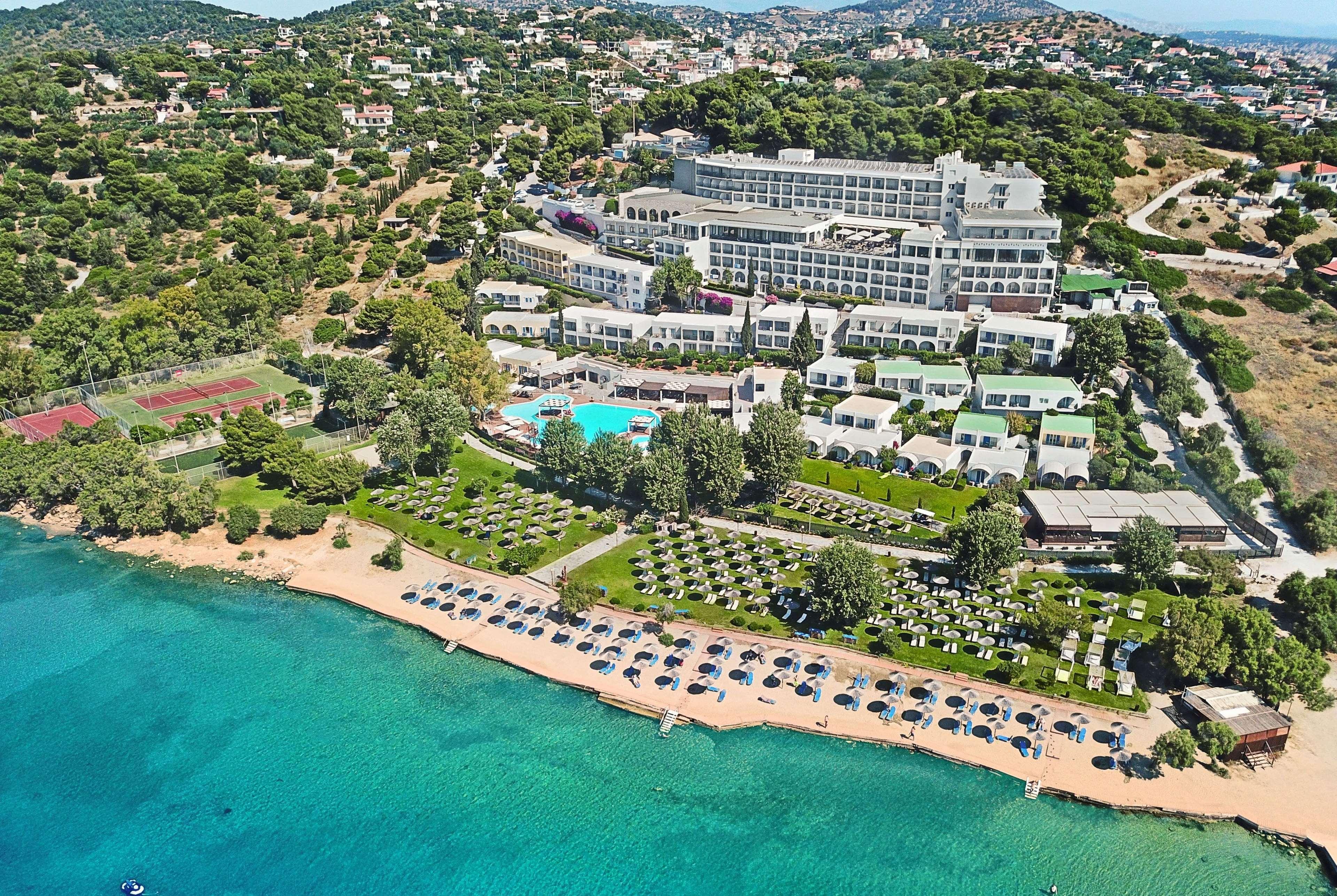 돌체 바이 윈덤 아티카 리비에라 호텔 브라브로나 외부 사진 The photo shows a scenic aerial view of a resort located near a beautiful coastline. The resort features a large building and various smaller accommodations. An inviting swimming pool is visible at the center, surrounded by sun loungers and greenery.